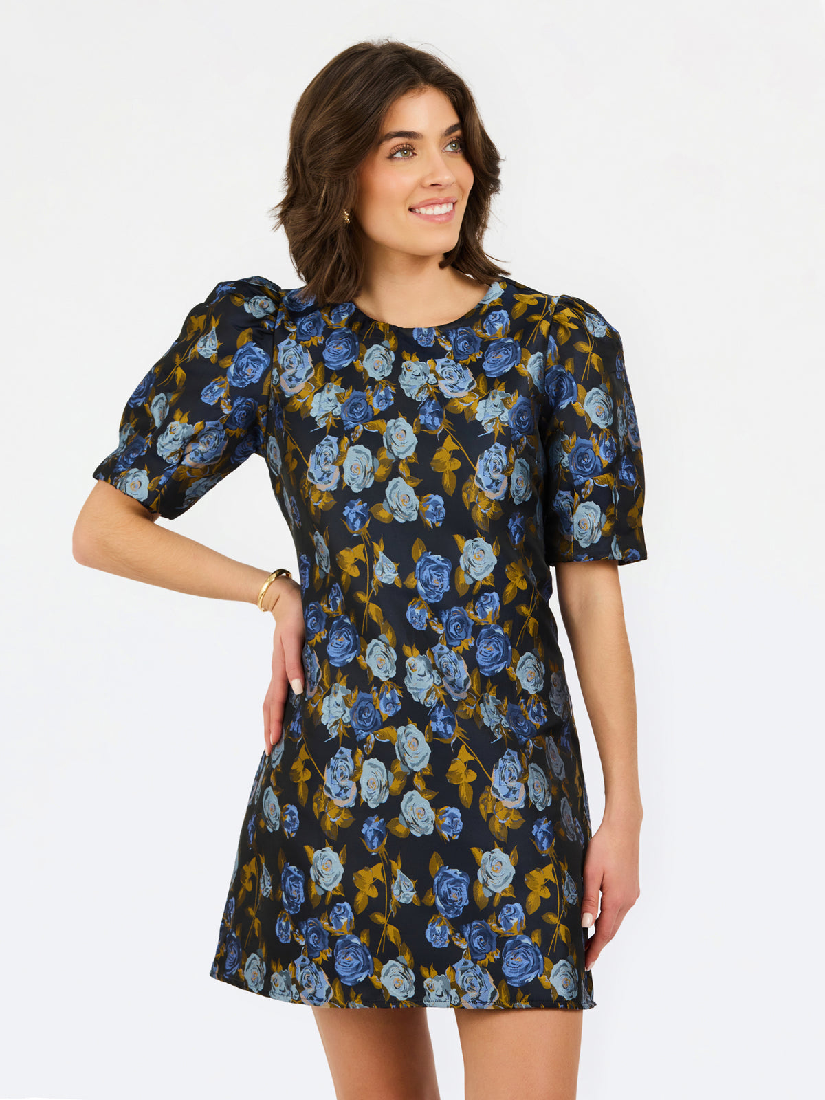 A woman modeling a short puff-sleeve dress with an all-over floral print in blue, light blue, and mustard yellow on a dark background, set against a plain white studio backdrop.