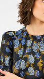 A close-up of a woman wearing a black blouse with a blue and light blue rose floral pattern and gold-toned leaves, paired with a gold earring.