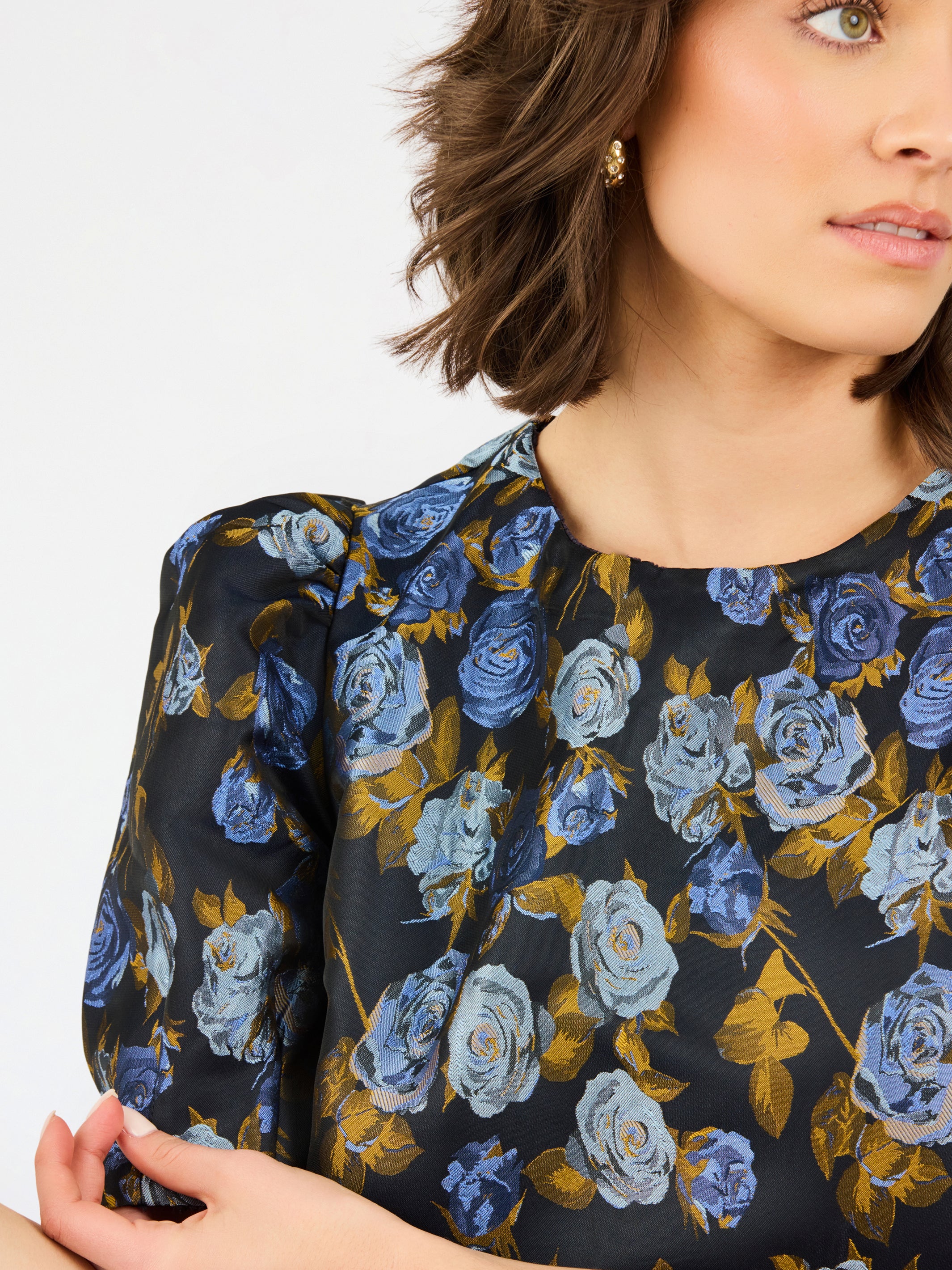 A close-up of a woman wearing a black blouse with a blue and light blue rose floral pattern and gold-toned leaves, paired with a gold earring.