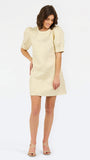 A woman modeling a short cream-colored textured dress with puff sleeves and a round neckline, paired with strappy high-heeled sandals, against a plain white studio background.