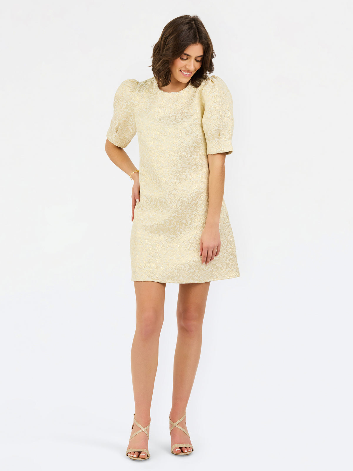 A woman modeling a short cream-colored textured dress with puff sleeves and a round neckline, paired with strappy high-heeled sandals, against a plain white studio background.