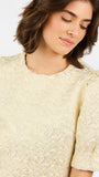 A close-up of a woman wearing a pale cream or light beige textured blouse with short puff sleeves and a round neckline, photographed against a plain light background.