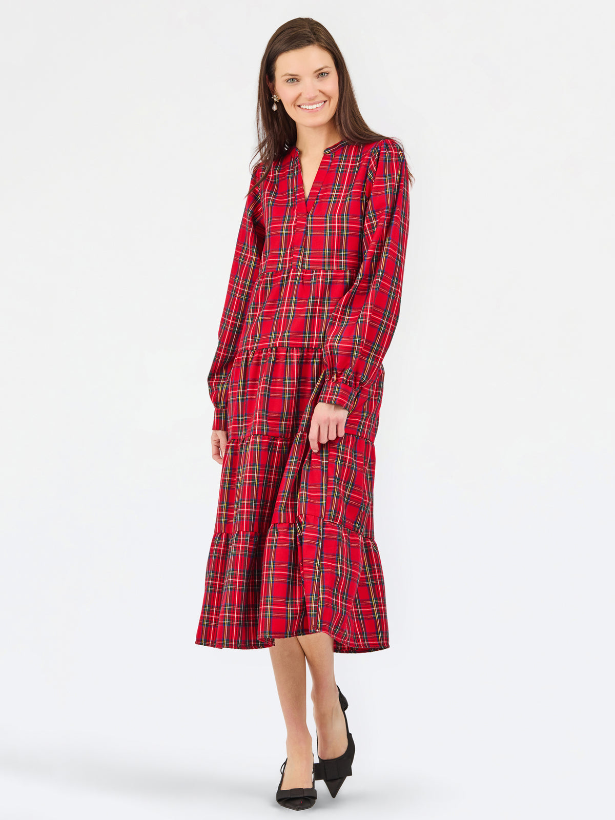 A woman modeling a red tartan plaid midi dress with long sleeves and a tiered skirt, paired with black heels, against a plain white studio background.
