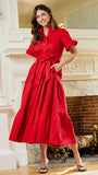 A woman modeling a bright red midi shirt dress with short sleeves, a button-up front, and a matching waist tie, standing in front of an ornate white fireplace in a sunlit room.