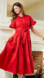 A woman wearing a bright red midi dress with short flutter sleeves, a buttoned collar, and a matching waist tie, standing in front of an ornate white fireplace in a grand interior.