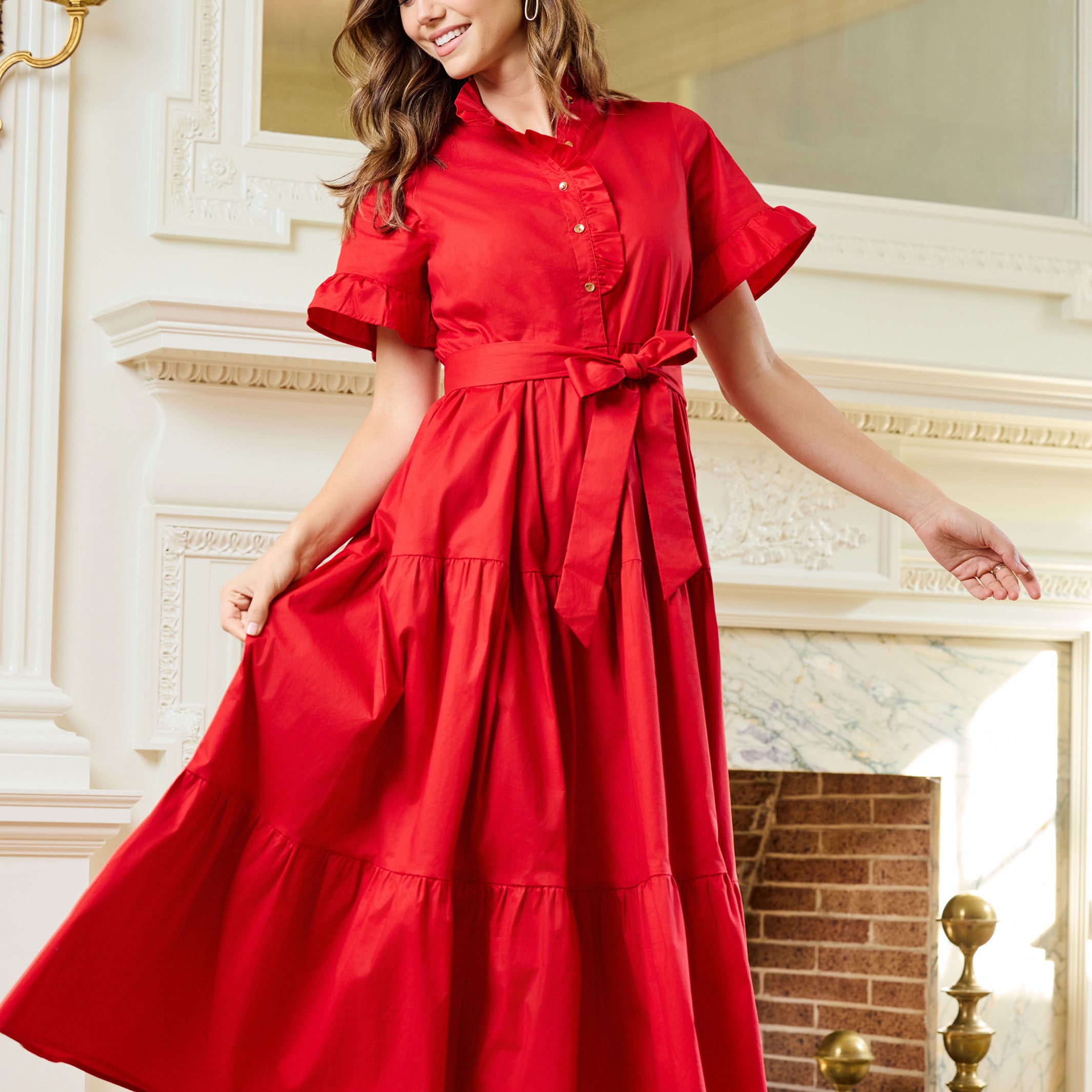 A woman wearing a bright red midi dress with short flutter sleeves, a buttoned collar, and a matching waist tie, standing in front of an ornate white fireplace in a grand interior.