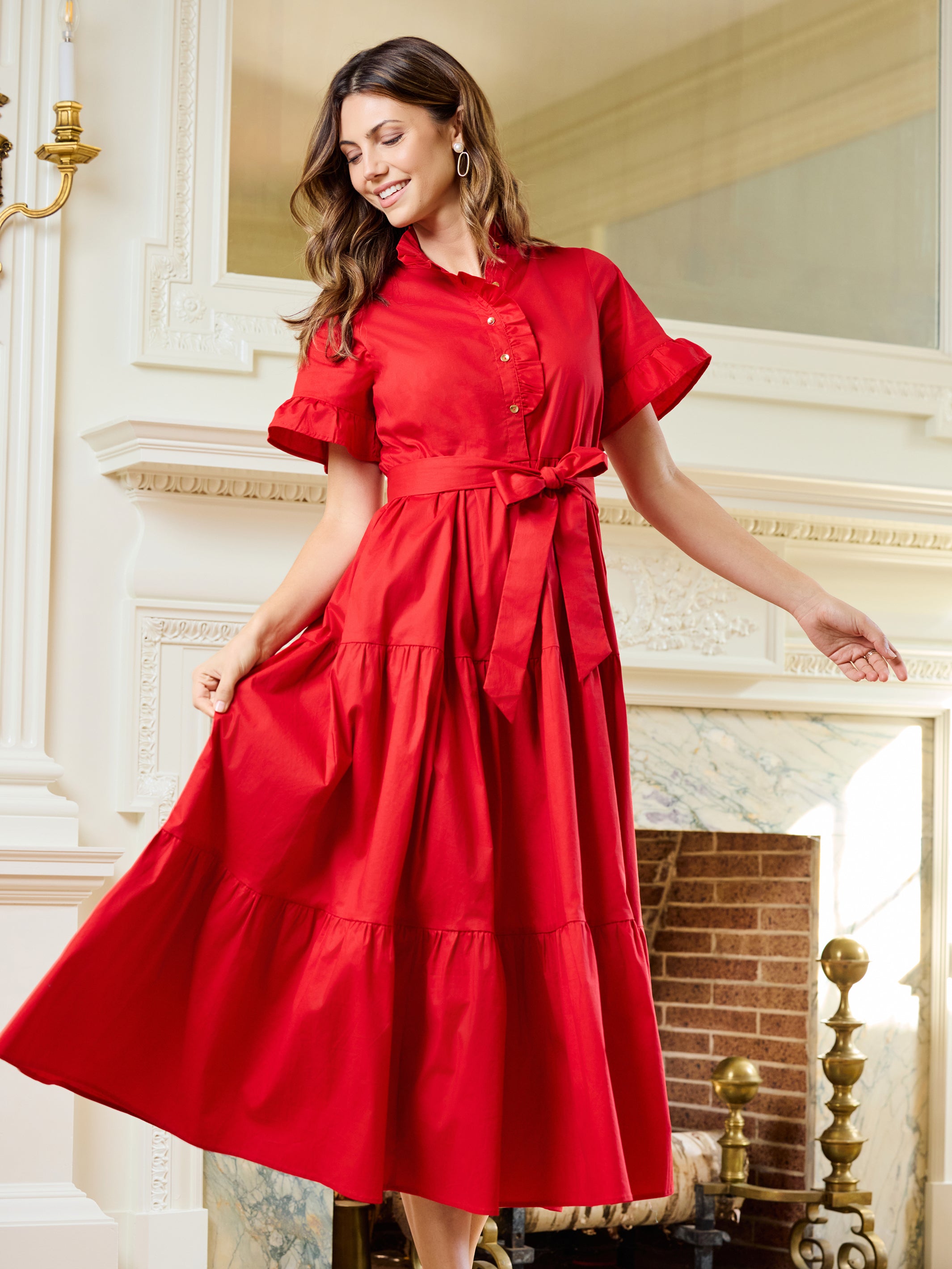 A woman wearing a bright red midi dress with short flutter sleeves, a buttoned collar, and a matching waist tie, standing in front of an ornate white fireplace in a grand interior.