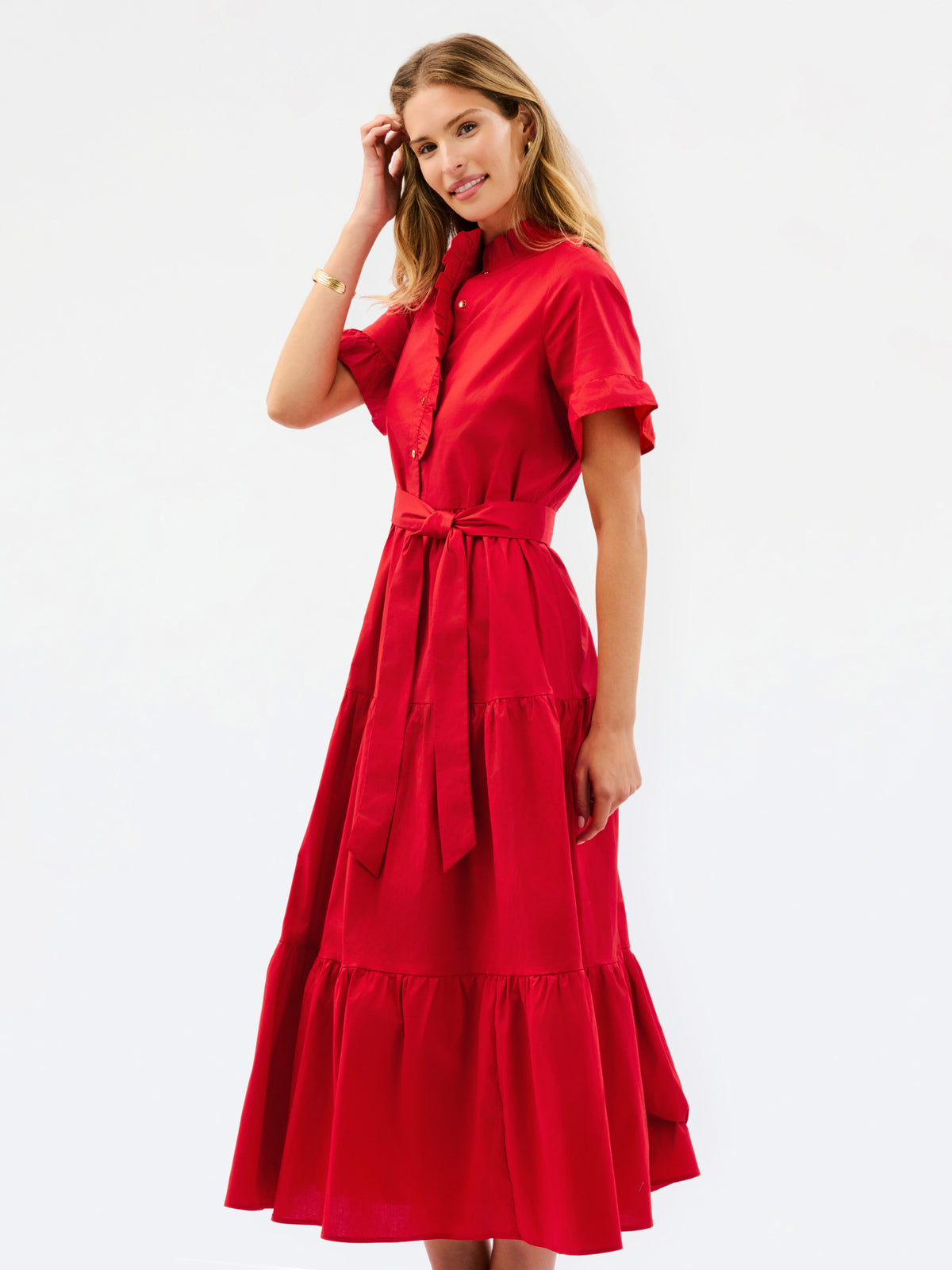 A woman modeling a bright red midi dress with short sleeves, a high collar, a button front, a matching waist tie, and a tiered skirt, photographed against a plain white studio background.