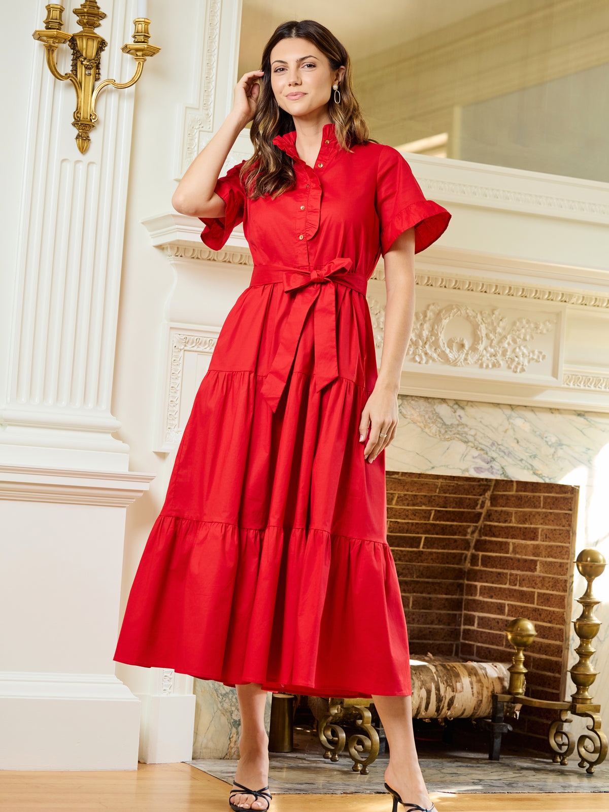 A woman modeling a bright red midi dress with short flutter sleeves, a button-front bodice, and a matching waist tie, posed in front of an ornate fireplace in a classical interior.