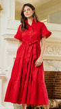 A woman wearing a bright red midi dress with short flutter sleeves, a button-up collar, and a matching waist tie, standing in front of an ornate white fireplace.