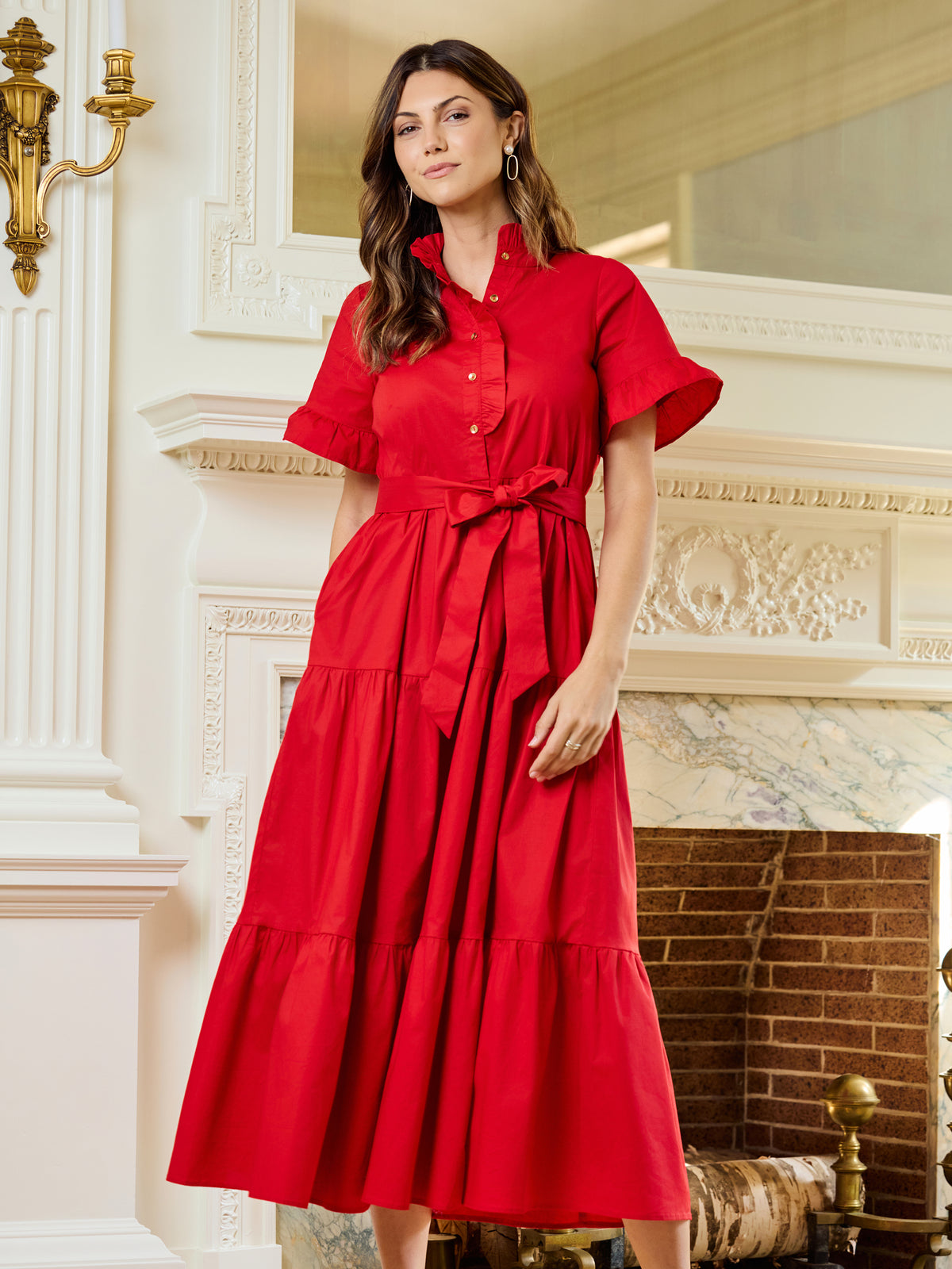 A woman wearing a bright red midi dress with short flutter sleeves, a button-up collar, and a matching waist tie, standing in front of an ornate white fireplace.
