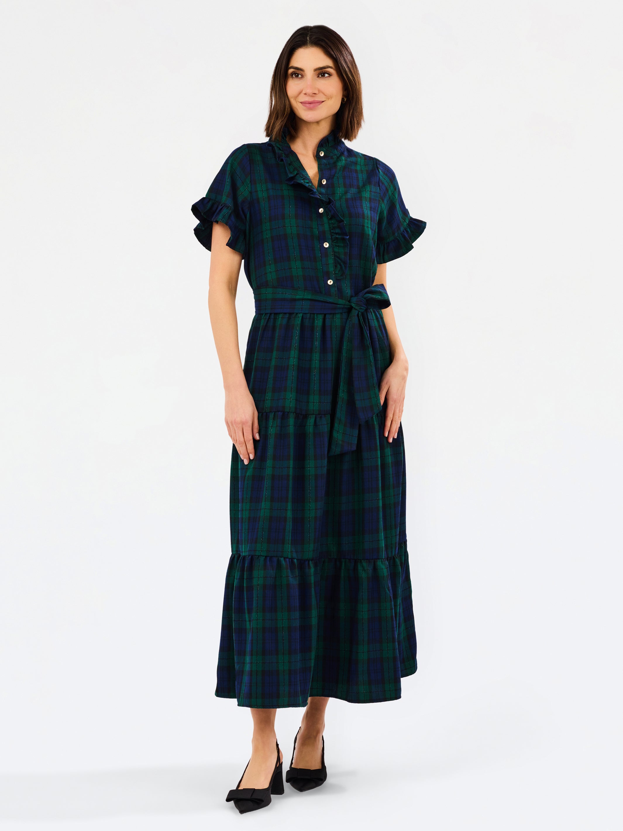 A woman modeling a dark green and black plaid midi dress with short flutter sleeves, a buttoned collar, and a matching waist tie, paired with black heels against a plain white studio background.