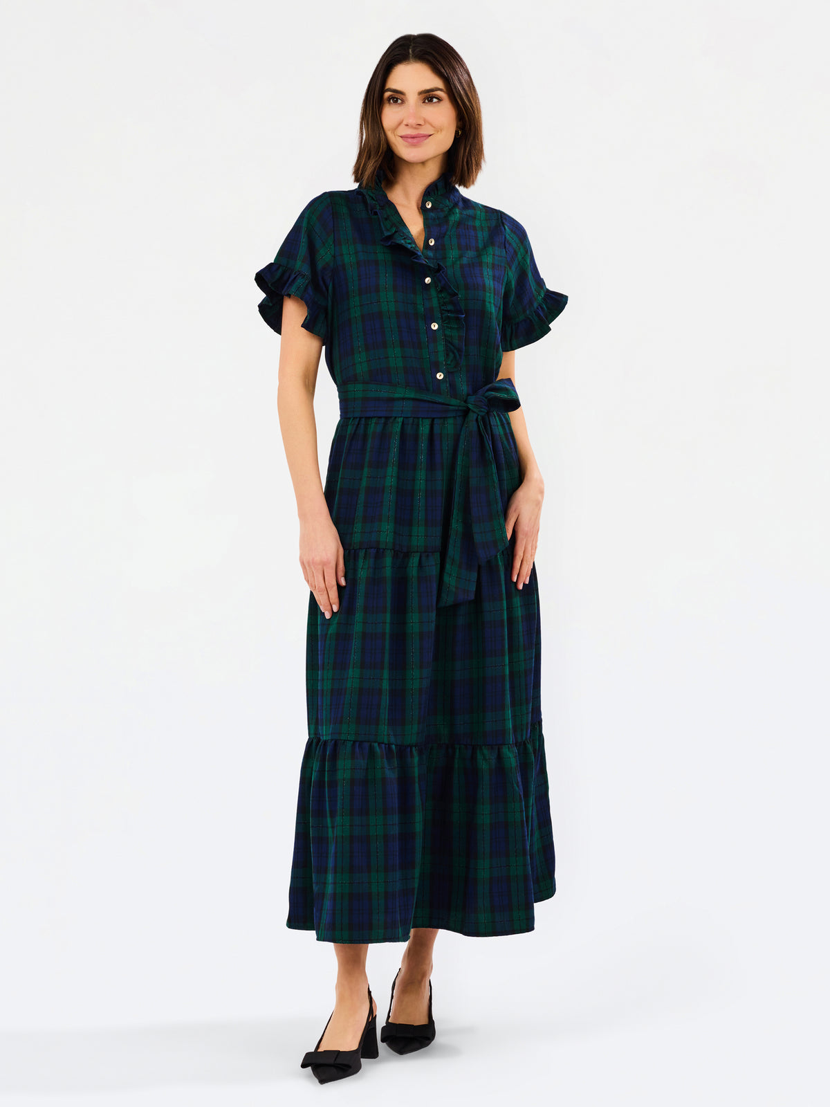 A woman modeling a dark green and black plaid midi dress with short flutter sleeves, a buttoned collar, and a matching waist tie, paired with black heels against a plain white studio background.