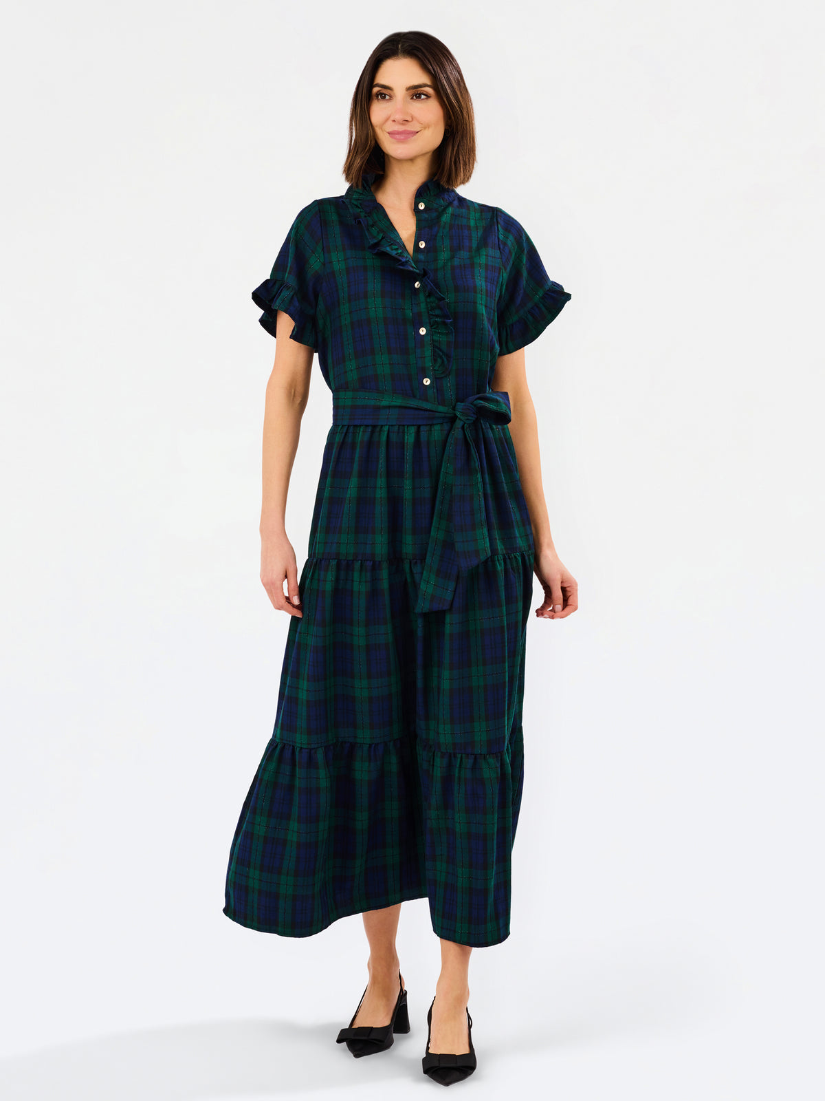 A woman modeling a dark green and black plaid midi shirt dress with short flutter sleeves, a button-up front, and a matching waist tie, paired with black heels against a plain white background.