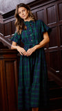 A woman wearing a dark green and navy plaid midi dress with flutter sleeves and a button-up front, standing on a dark wood staircase.