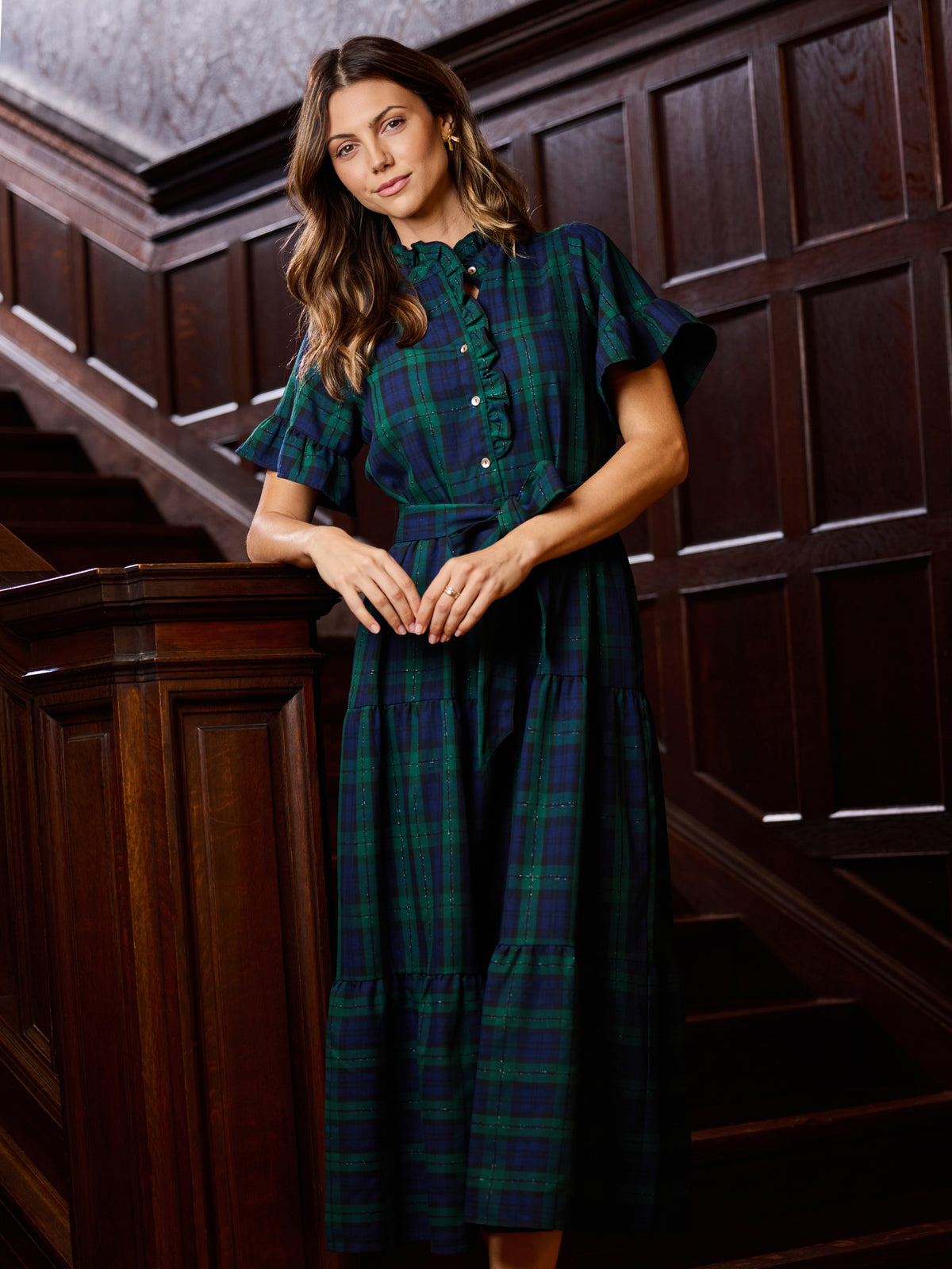 A woman wearing a dark green and navy plaid midi dress with flutter sleeves and a button-up front, standing on a dark wood staircase.