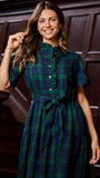 A woman wearing a dark green and navy plaid short-sleeve dress with a button-up front and waist tie, standing in front of a dark wood paneled wall.