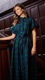 A woman wearing a dark green and navy plaid midi dress with short flutter sleeves, a buttoned high collar, and a matching waist tie, posing in front of a dark wood-paneled staircase.