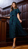 A woman wearing a dark green and navy plaid midi dress with flutter sleeves and a tie waist, posing on a wooden staircase.