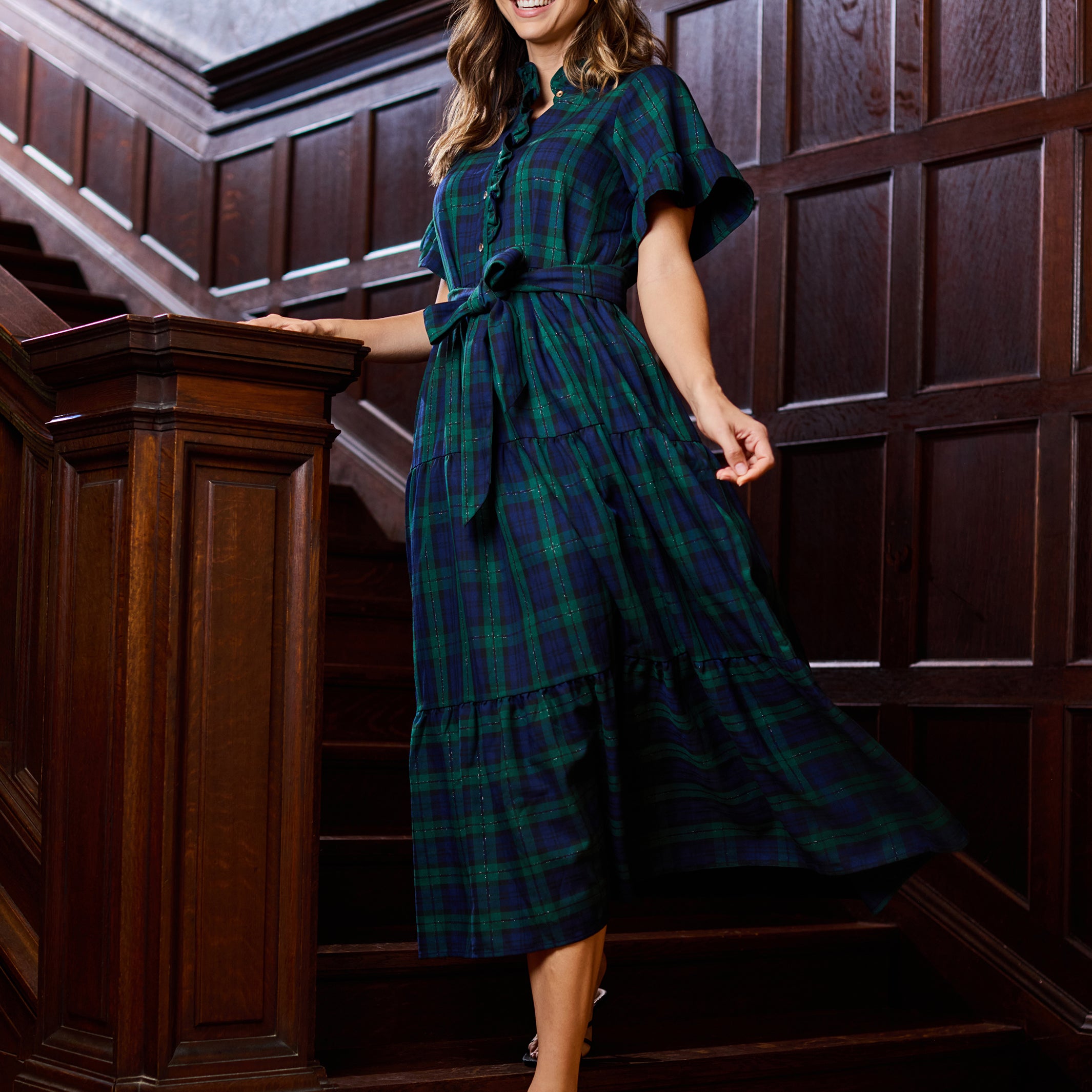 A woman wearing a dark green and navy plaid midi dress with flutter sleeves and a tie waist, posing on a wooden staircase.