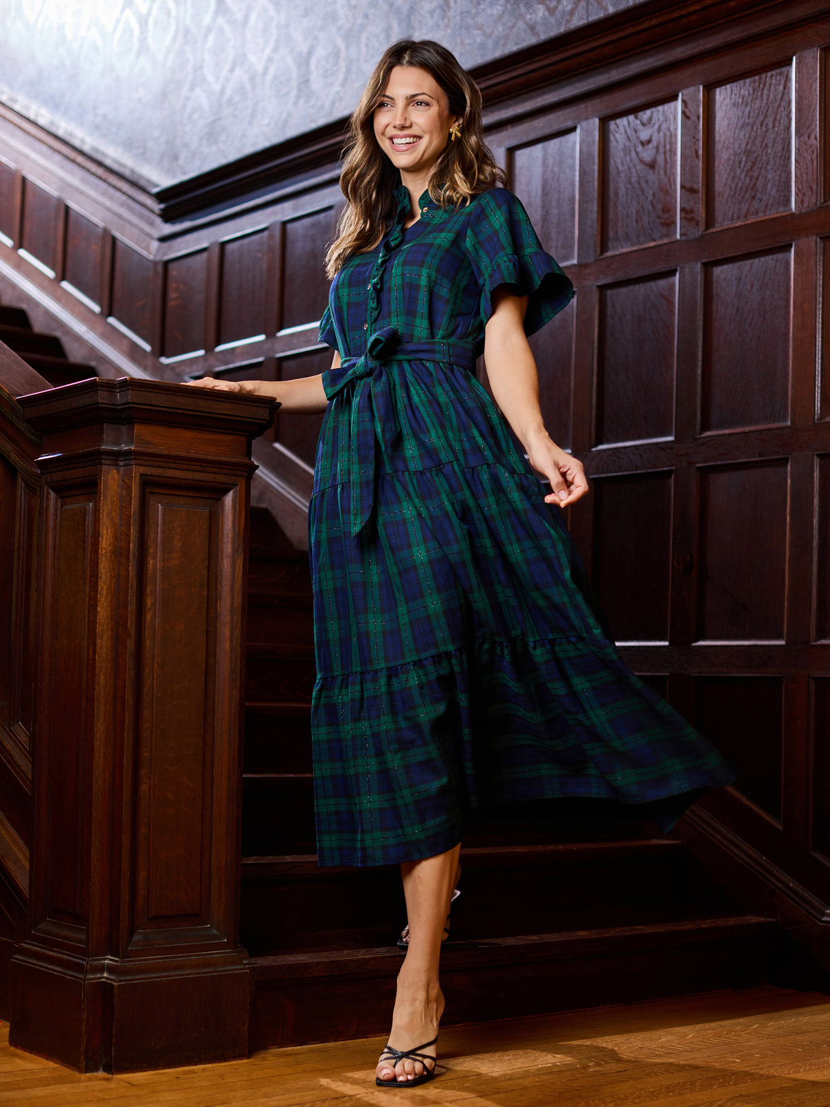 A woman wearing a dark green and navy plaid midi dress with flutter sleeves and a tie waist, posing on a wooden staircase.