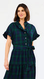 A woman wearing a dark green and navy plaid shirt dress with short flutter sleeves, a buttoned collar, and a matching tie belt at the waist, photographed against a plain light gray studio background.