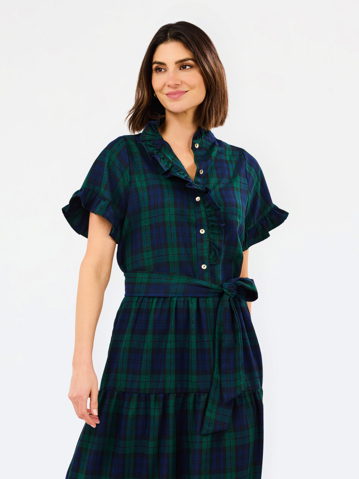 A woman wearing a dark green and navy plaid shirt dress with short flutter sleeves, a buttoned collar, and a matching tie belt at the waist, photographed against a plain light gray studio background.