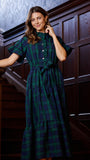 A woman wearing a green and navy plaid midi dress with short flutter sleeves, a buttoned high neck, and a matching waist tie, posing on a dark wood staircase.