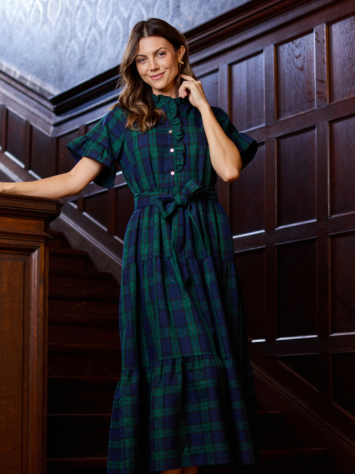 A woman wearing a green and navy plaid midi dress with short flutter sleeves, a buttoned high neck, and a matching waist tie, posing on a dark wood staircase.