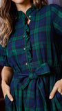 A close-up of a woman wearing a dark green and navy plaid shirt dress with short puff sleeves, a ruffled collar, and a matching fabric tie belt at the waist.