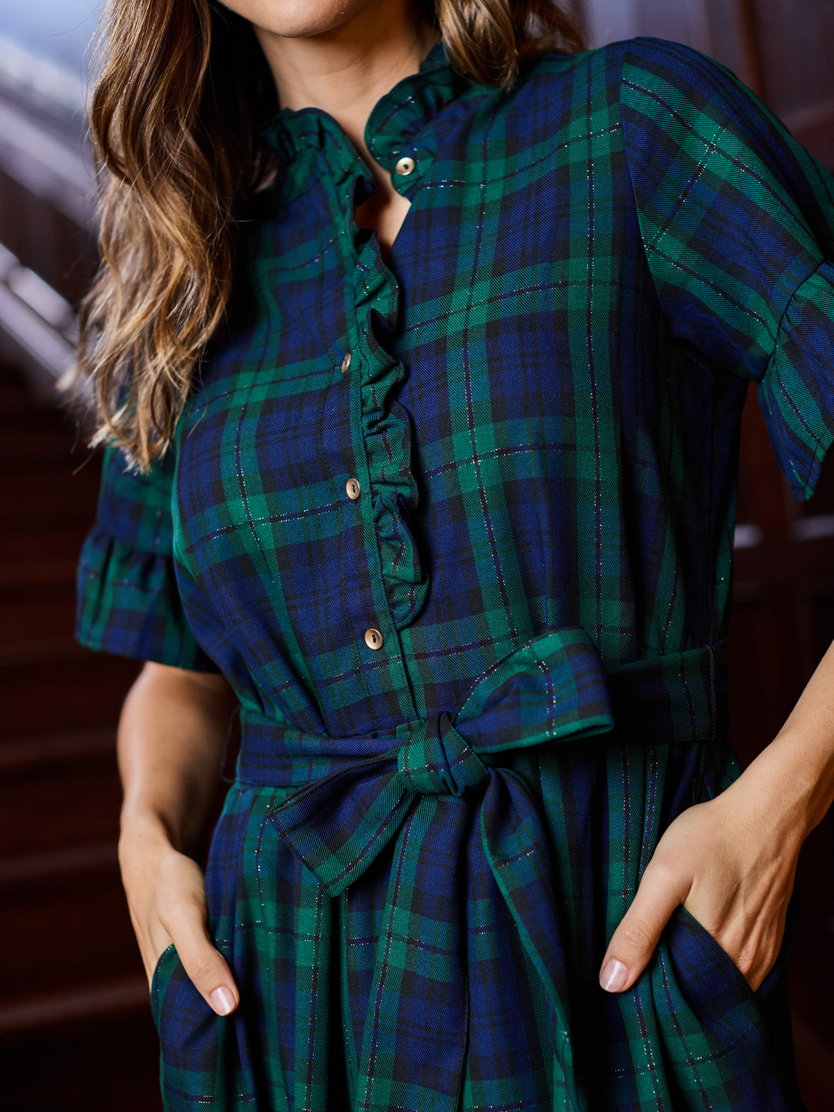 A close-up of a woman wearing a dark green and navy plaid shirt dress with short puff sleeves, a ruffled collar, and a matching fabric tie belt at the waist.