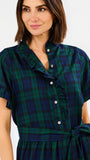 A woman wearing a dark green and navy plaid shirt dress with ruffled trim, a buttoned collar, and a tie waist, shown against a plain light gray background.