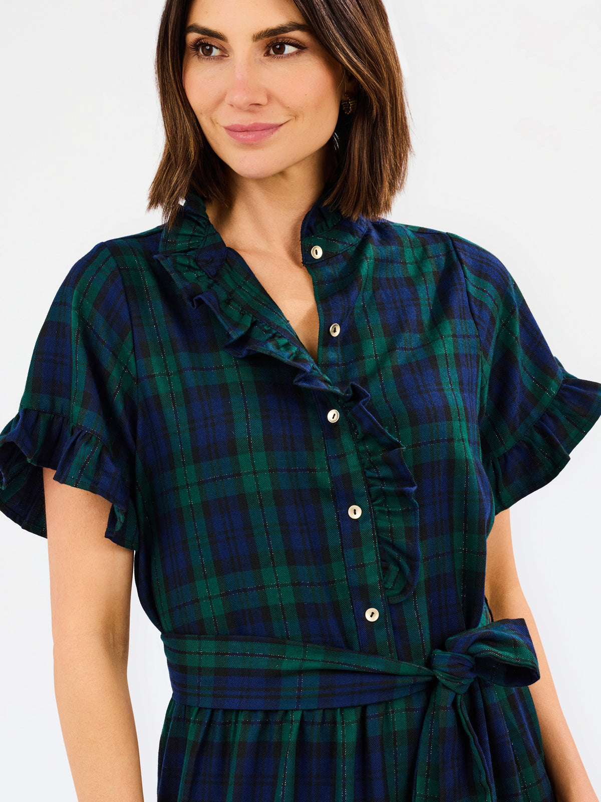 A woman wearing a dark green and navy plaid shirt dress with ruffled trim, a buttoned collar, and a tie waist, shown against a plain light gray background.
