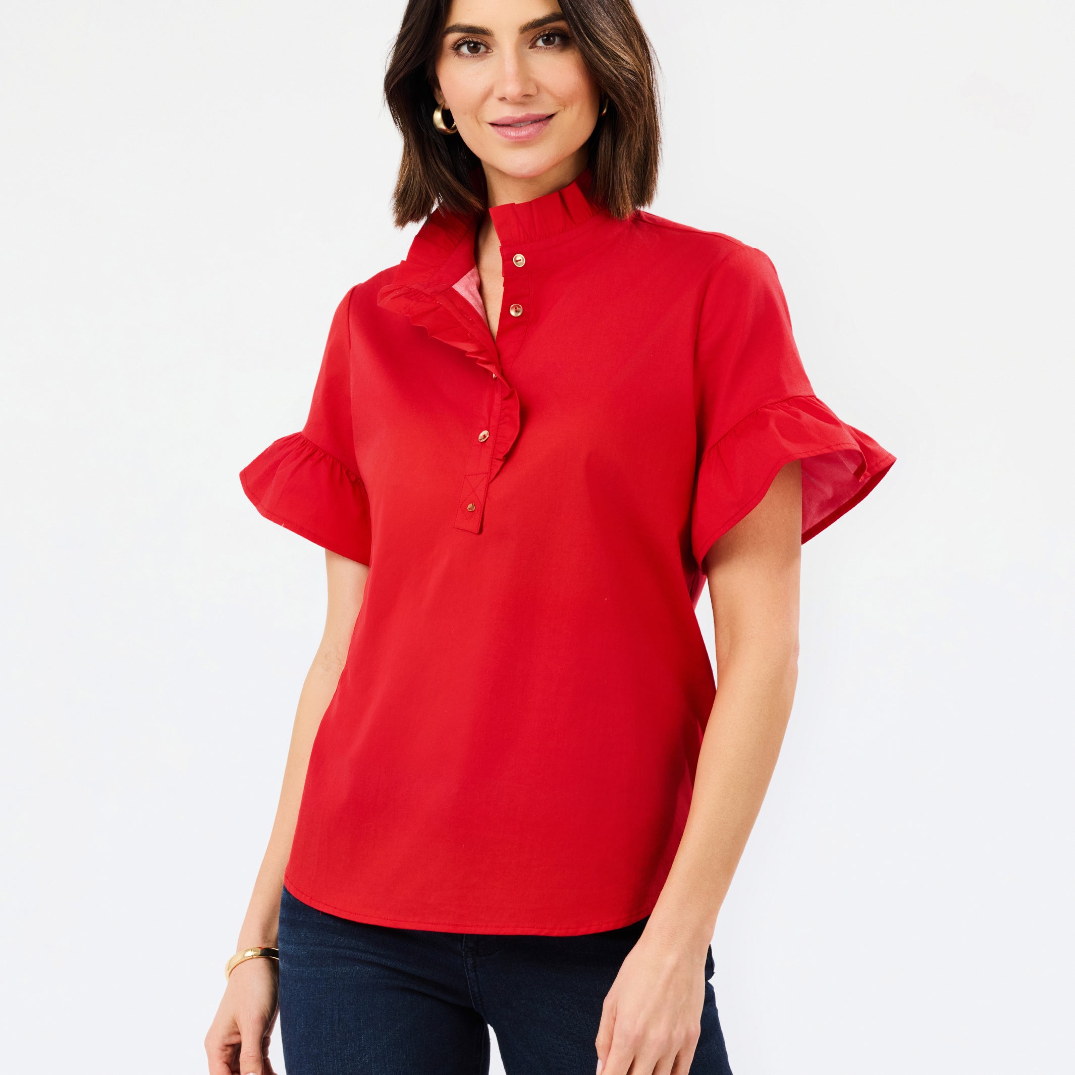 A woman modeling a bright red short-sleeve blouse with a high ruffled collar, button placket, and flutter sleeves, paired with dark blue jeans against a plain white studio background.