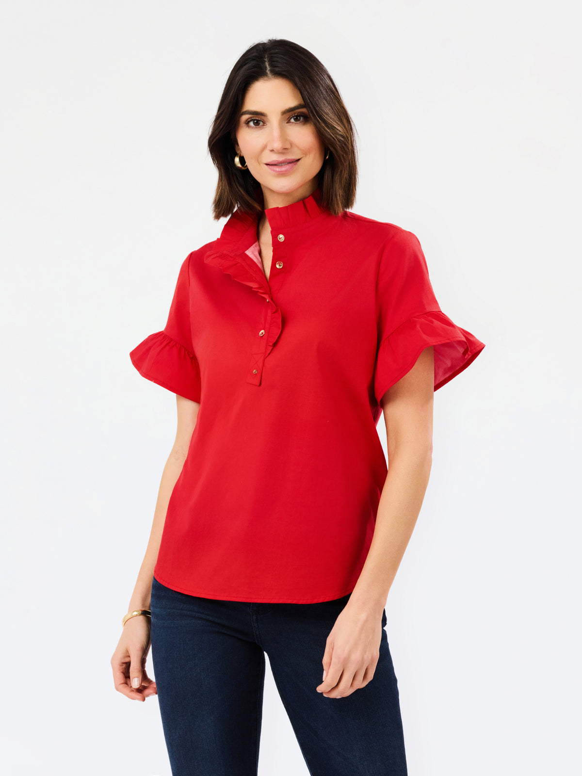 A woman modeling a bright red short-sleeve blouse with a high ruffled collar, button placket, and flutter sleeves, paired with dark blue jeans against a plain white studio background.