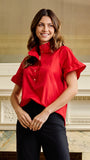 A woman wearing a vibrant red short-sleeve blouse with a high mock neck and ruffled sleeves, paired with black pants, standing in front of a classic cream-colored fireplace.