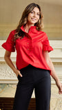 A woman wearing a bright red short-sleeve blouse with a high ruffled collar and black pants, standing in front of an ornate indoor fireplace.