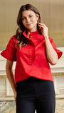 A woman wearing a bright red short-sleeve blouse with a ruffled high collar and button placket, paired with black pants, standing in front of an elegant cream-colored wall with classical molding.