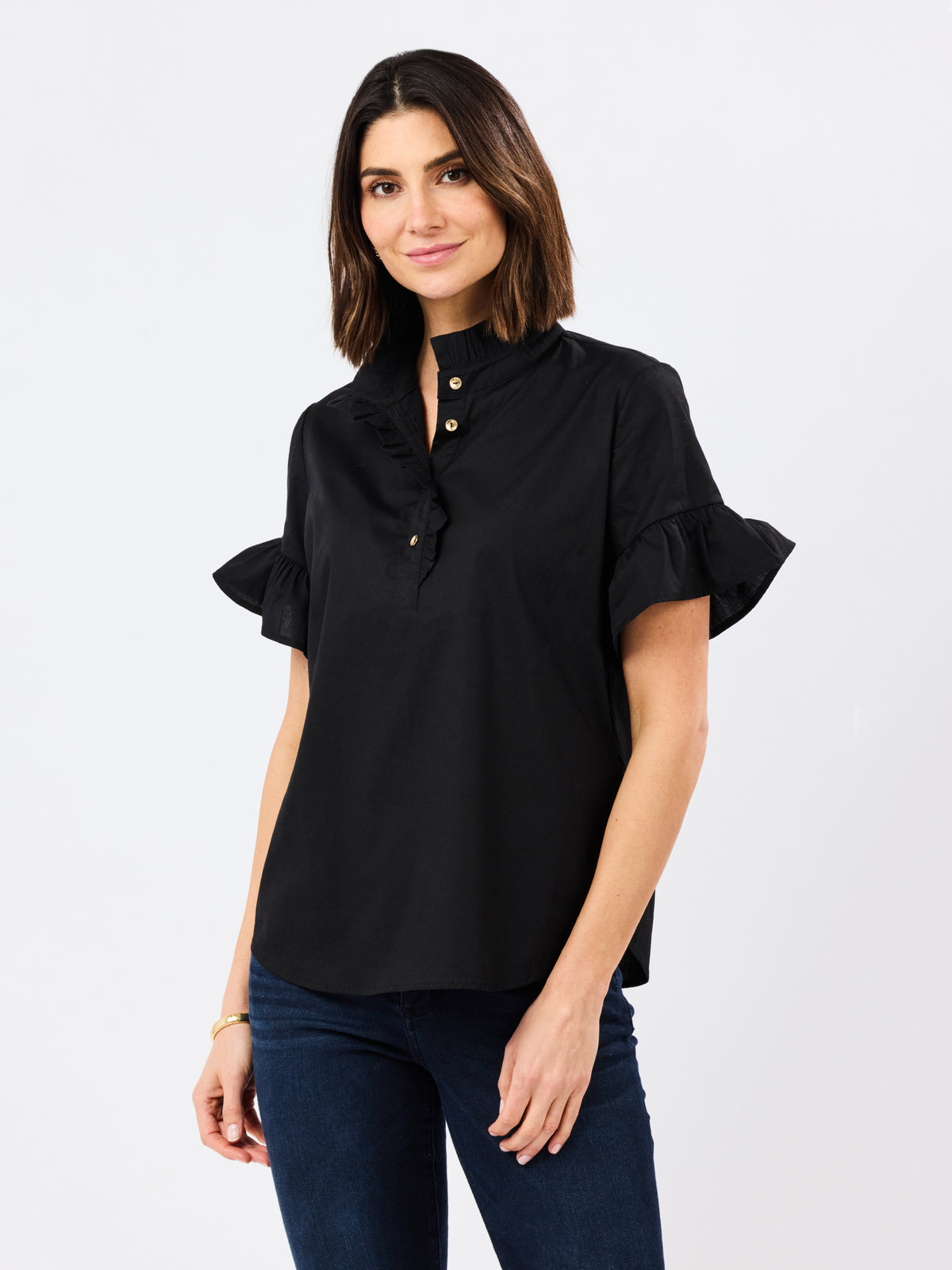 A woman modeling a black short-sleeve blouse with ruffled trim and gold buttons, paired with dark blue jeans, against a plain light gray studio background.