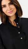 A close-up of a woman wearing a black blouse with gold buttons and a ruffled collar, set against a plain light gray background.