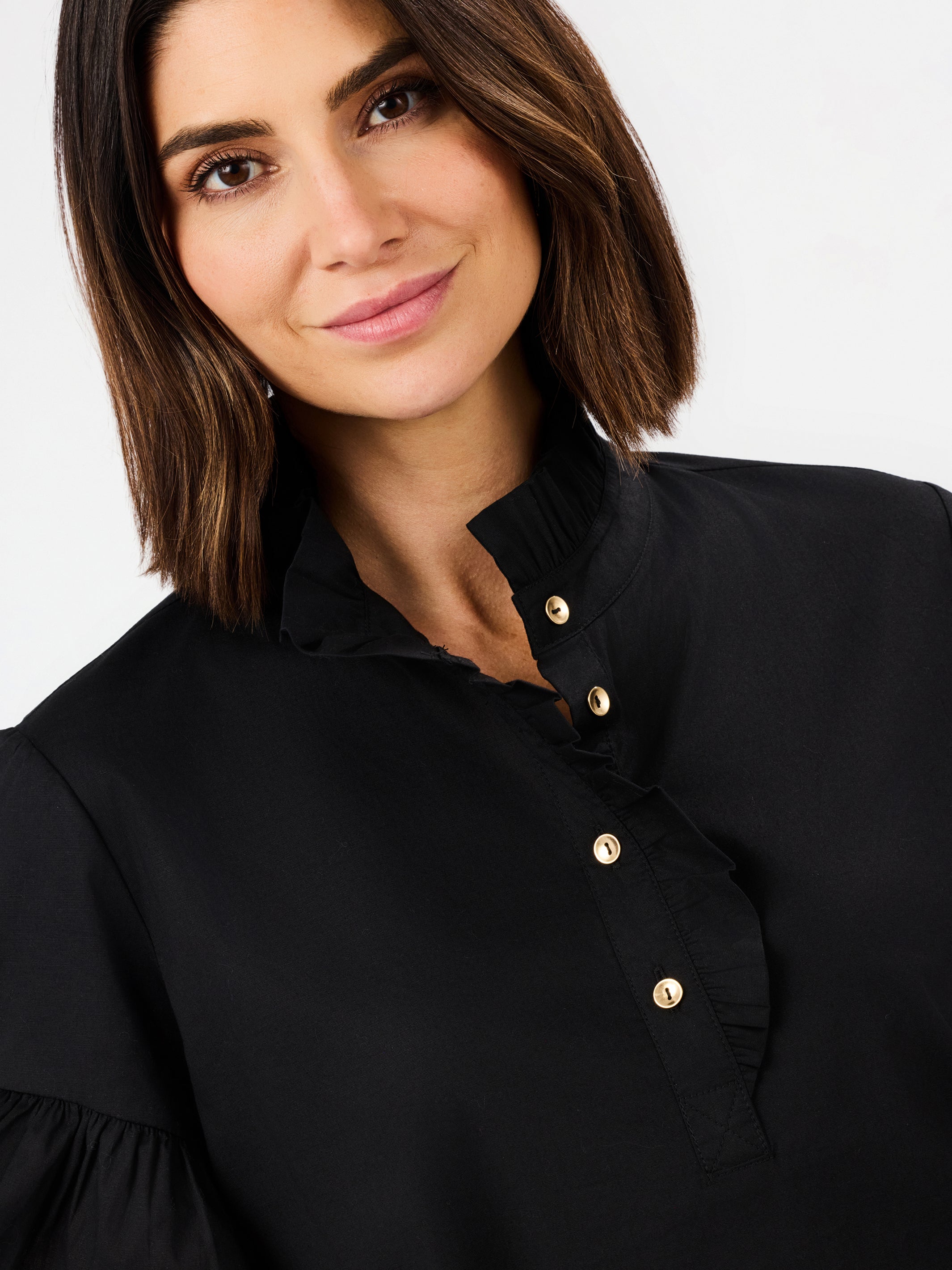 A close-up of a woman wearing a black blouse with gold buttons and a ruffled collar, set against a plain light gray background.