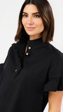 A woman wearing a black short-sleeve blouse with a high ruffled collar, gold buttons, and ruffled cuffs, photographed against a plain light gray studio background.