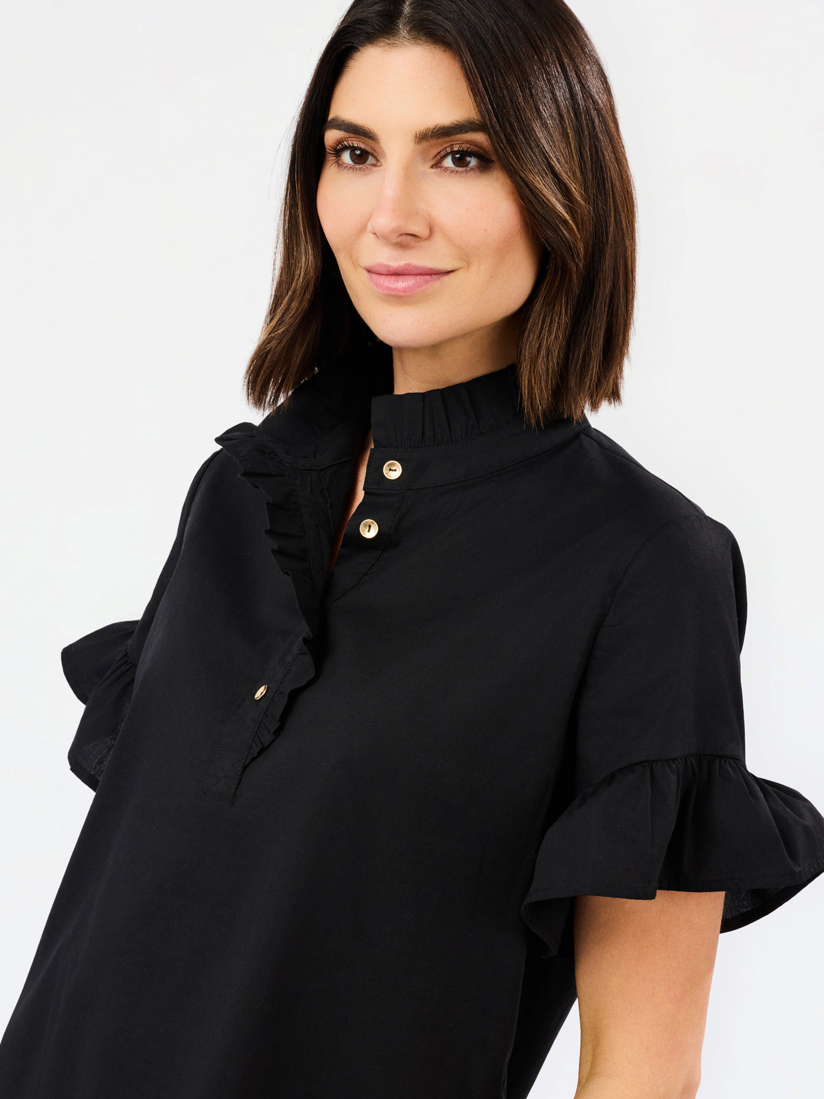 A woman wearing a black short-sleeve blouse with a high ruffled collar, gold buttons, and ruffled cuffs, photographed against a plain light gray studio background.