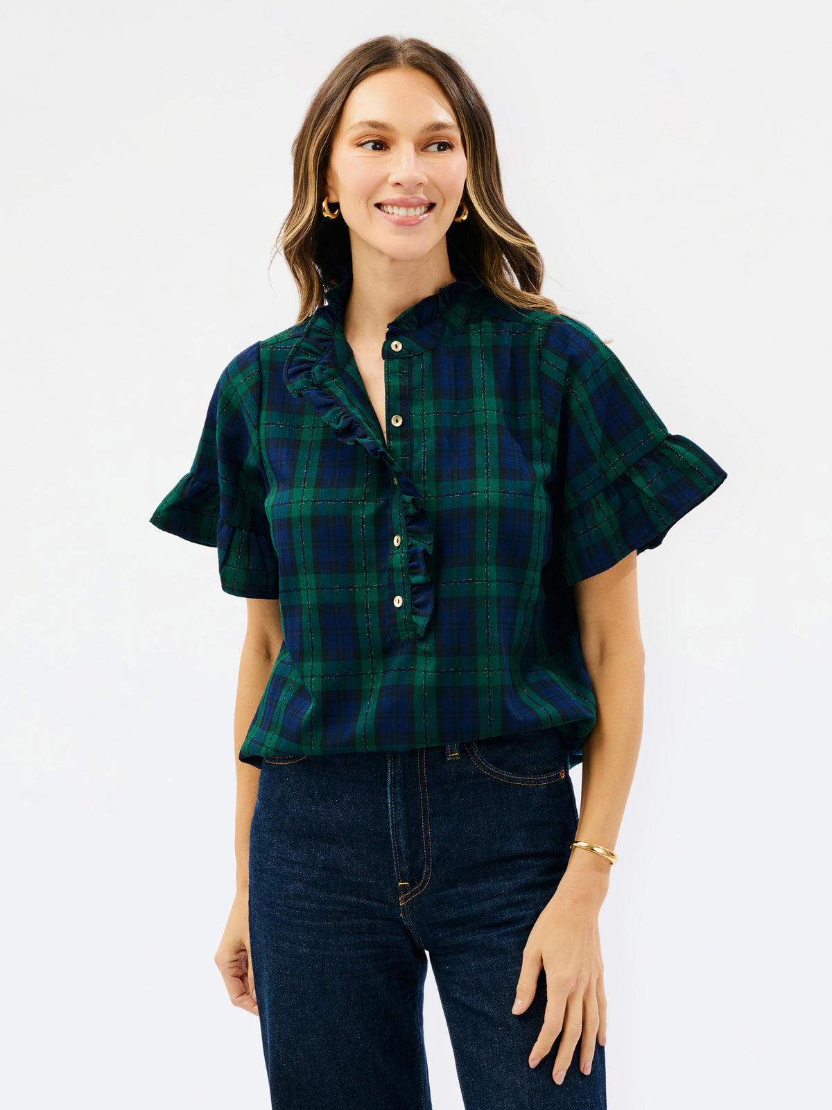 A woman modeling a short-sleeve plaid blouse with ruffle trim and button-front detail, paired with dark blue jeans, against a plain white studio background.
