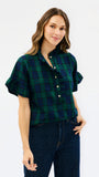 A woman modeling a short-sleeve button-front plaid shirt in dark green and navy with cream buttons, paired with dark blue jeans, against a plain white studio background.