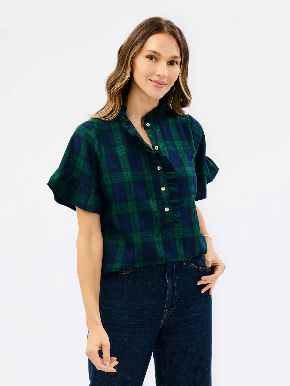 A woman modeling a short-sleeve button-front plaid shirt in dark green and navy with cream buttons, paired with dark blue jeans, against a plain white studio background.