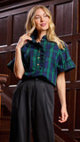 A woman models a short-sleeve button-up shirt with a green and navy plaid pattern and ruffled trim, paired with black wide-leg trousers, set against a dark wood-paneled background.