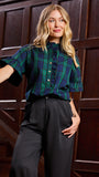 A woman posing in front of a dark wood paneled wall, wearing a green and blue plaid short-sleeve blouse with black ruffle trim and black trousers.
