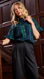 A woman wearing a green and navy plaid short-sleeve button-up shirt with ruffled cuffs and dark wide-leg trousers, standing in front of a dark wood-paneled wall.