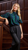 A blonde woman leans against a dark wood staircase railing, wearing a green and navy plaid short-sleeve button-up shirt with dark charcoal trousers.